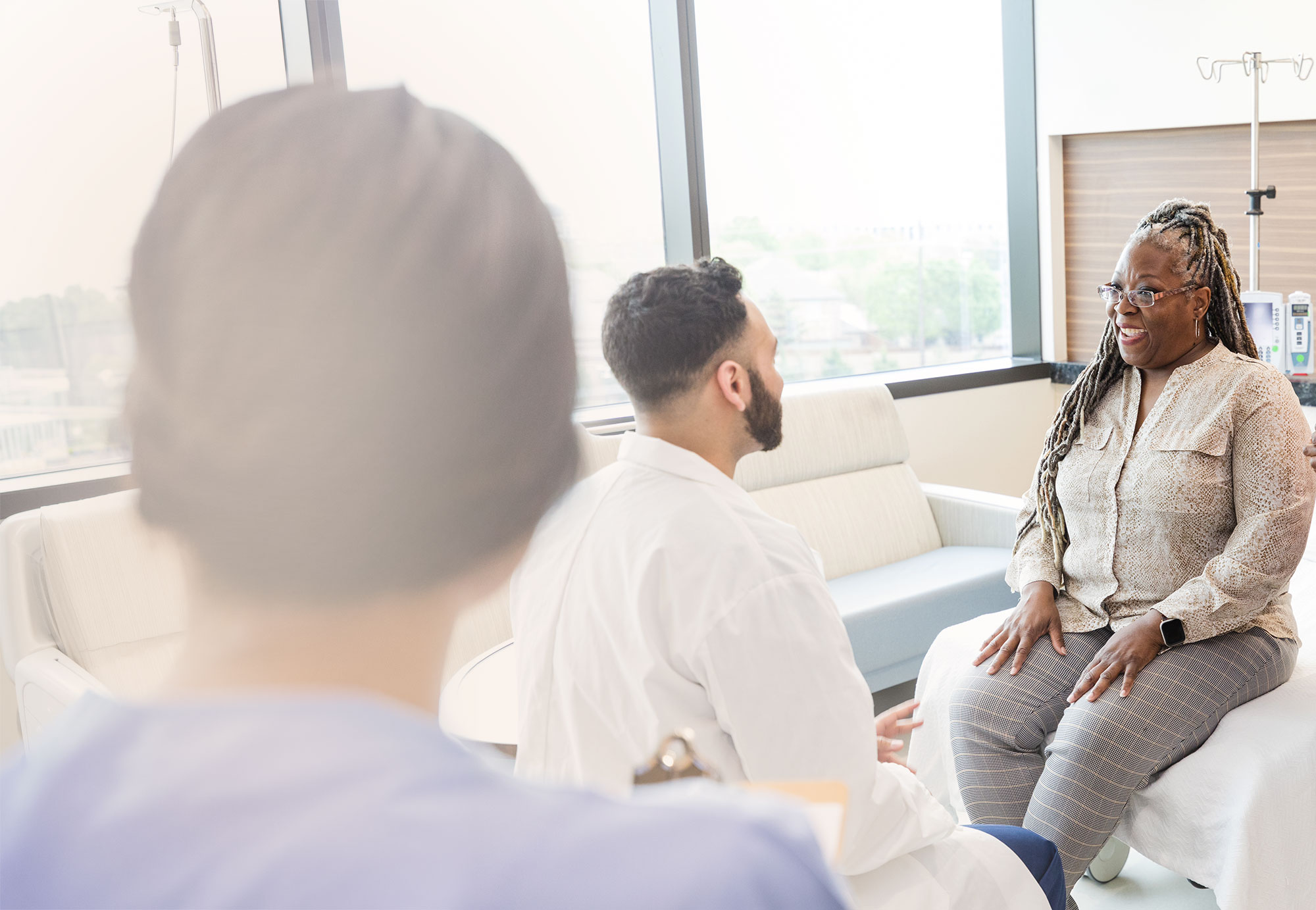 A patient speaks with two healthcare professionals in a bright exam room.