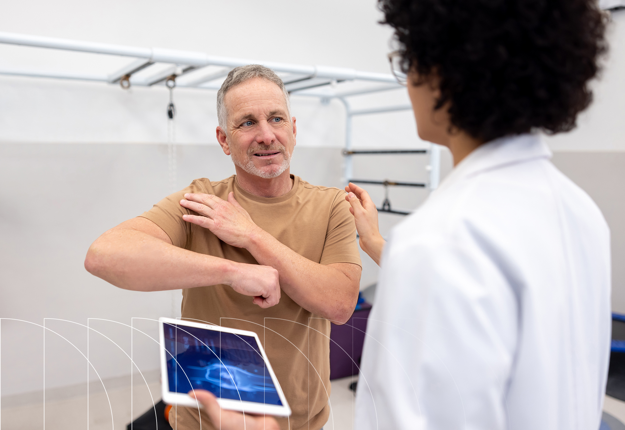 An older male patient points to his shoulder while speaking with a healthcare professional in a clinic.