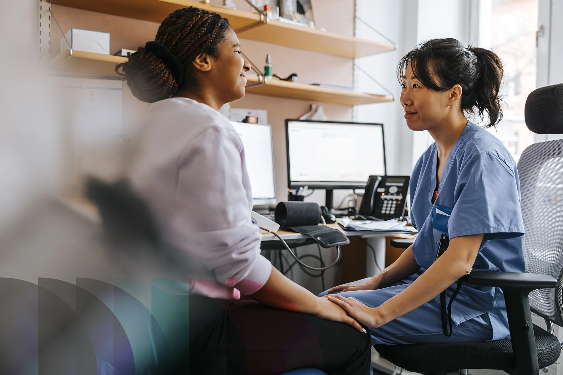 A medical professional sits with a patient in an exam room while talking and holding the patient’s hands.