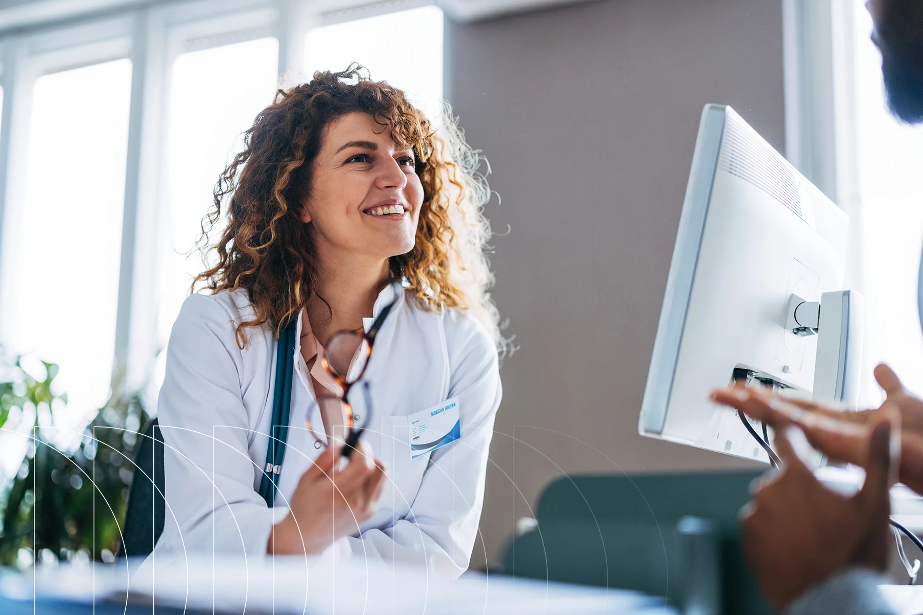 A doctor smiles while speaking with a patient at a computer in a medical office.