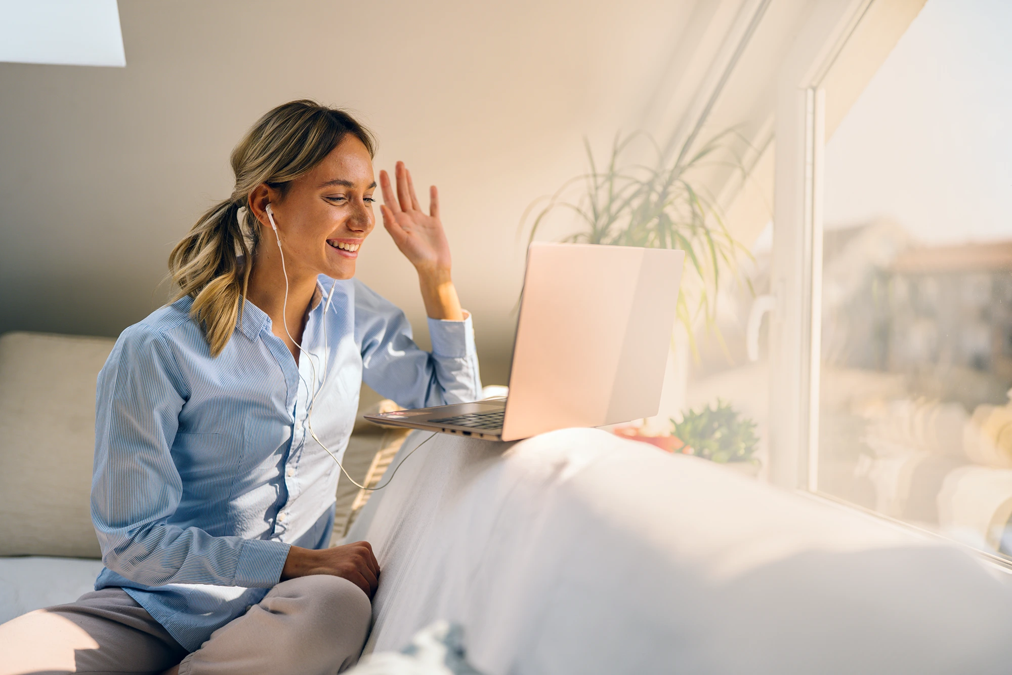A young woman sitting on a couch in a brightly lit room wears ear buds and engages in a virtual conversation on her laptop.