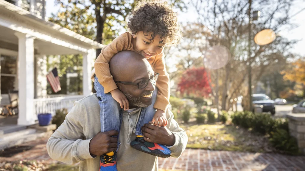 Father carrying toddler boy on shoulders in front of home
