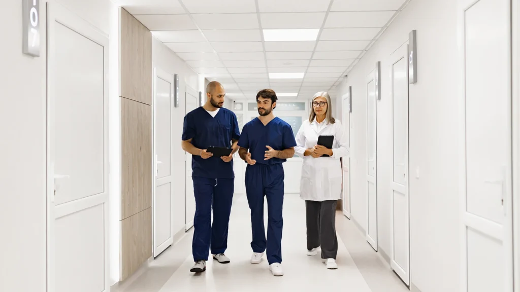 Three medical professionals, including a doctor, walk down a bright hospital corridor, engaged in conversation.